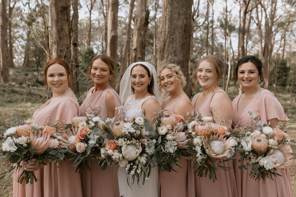 Bridesmaids in mauve dresses hold elegant white and pink floral bouquets while standing outdoors.