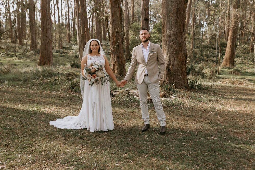 A couple in wedding attire holds hands while standing in a serene wooded forest setting.