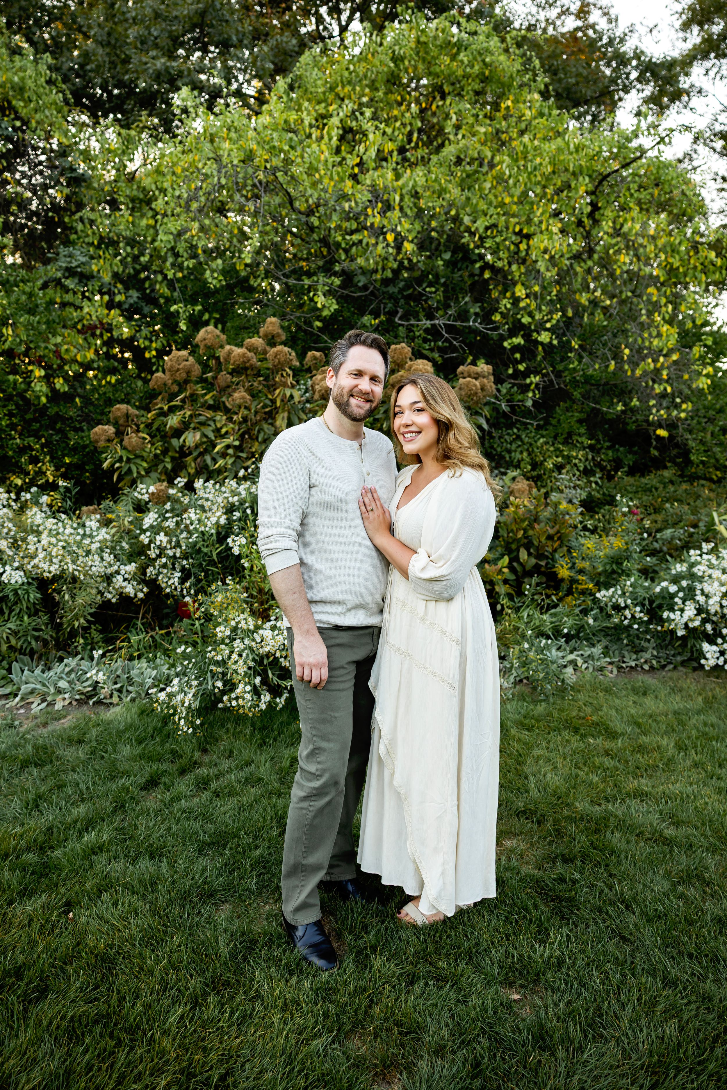 A couple in white and grey outfits poses together on a grassy hillside with flowering bushes in the background.