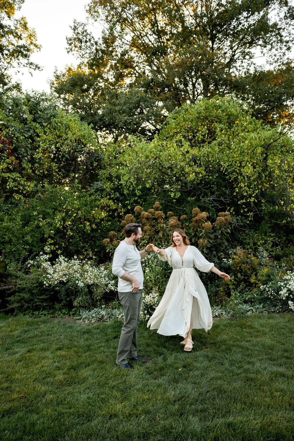 A couple shares a romantic dance together on a grassy hillside surrounded by lush green trees and bushes at sunset.