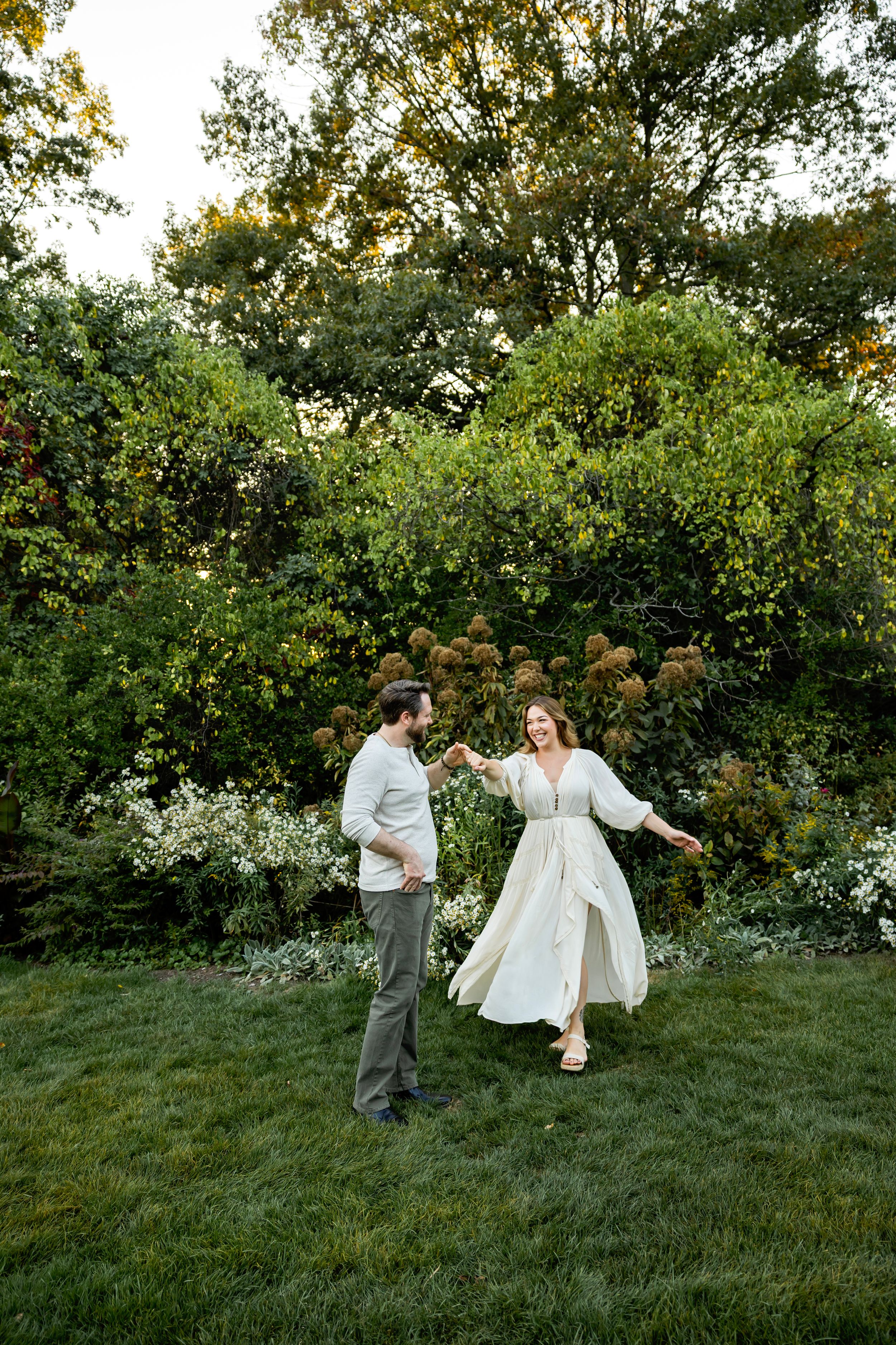 A couple shares a romantic dance together on a grassy hillside surrounded by lush green trees and bushes at sunset.