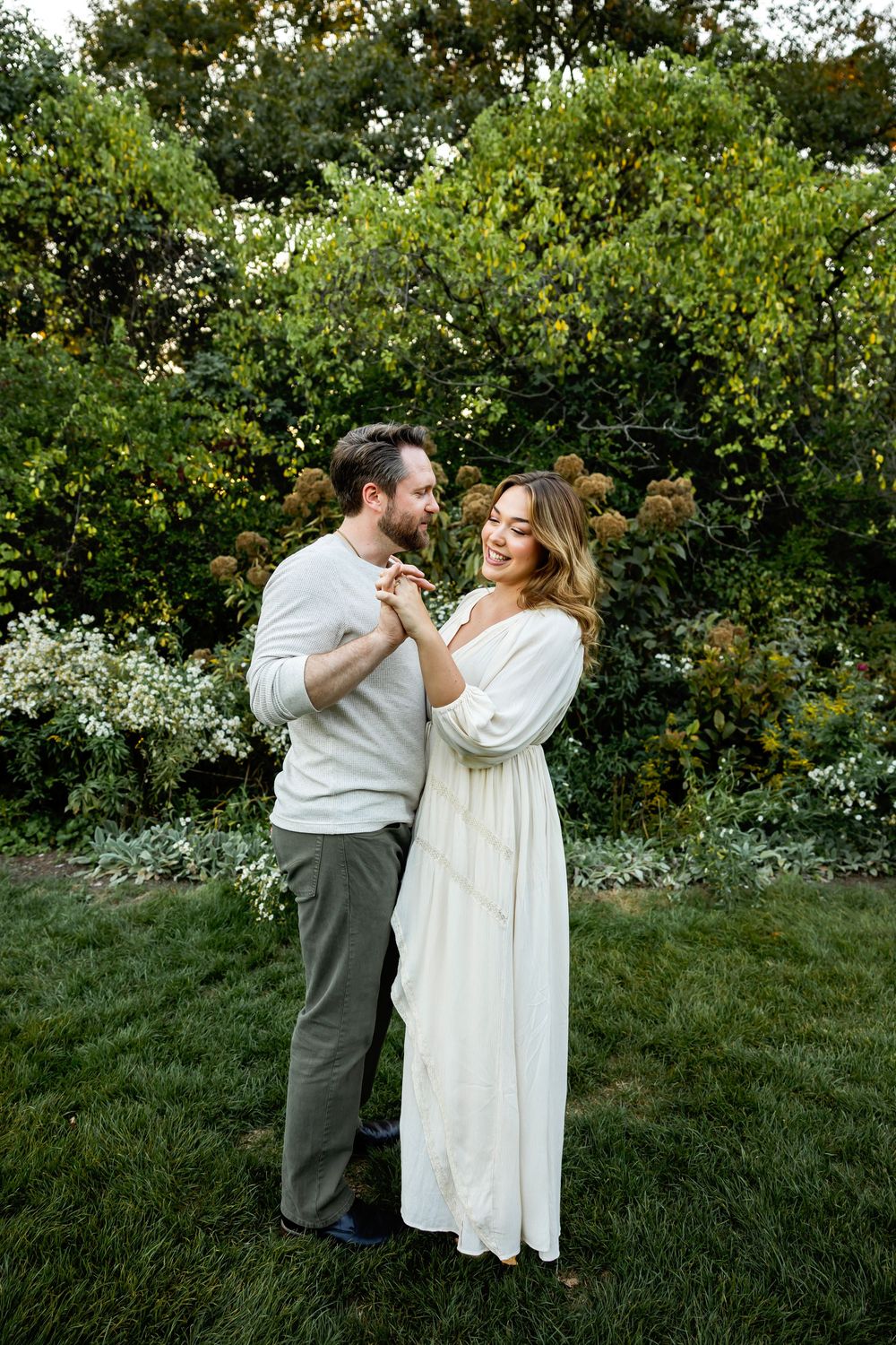 A couple shares a romantic moment on a lush green lawn with blooming trees and bushes in the background.