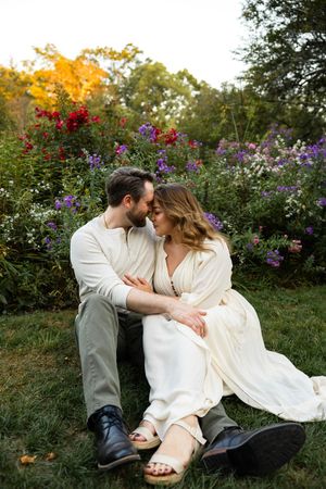 Couple in light colored clothing embraces on grass surrounded by colorful garden flowers at sunset.