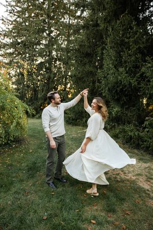 A couple dances joyfully together on a grassy hillside at sunset, the bride's white dress flowing elegantly in motion.