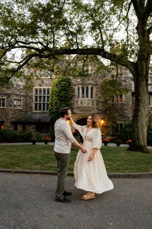 A couple shares a romantic moment on a driveway in front of a grand stone mansion with Gothic architecture at dusk.