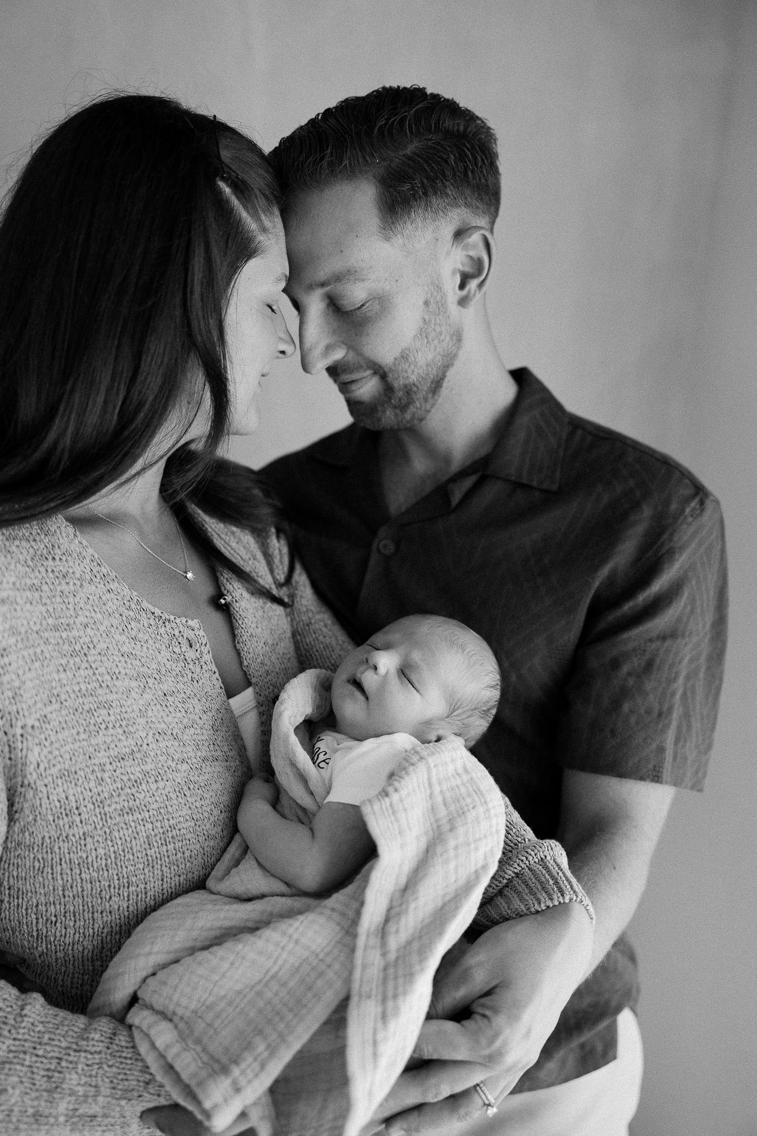A tender black and white portrait of parents sharing an intimate moment while holding their sleeping newborn baby.