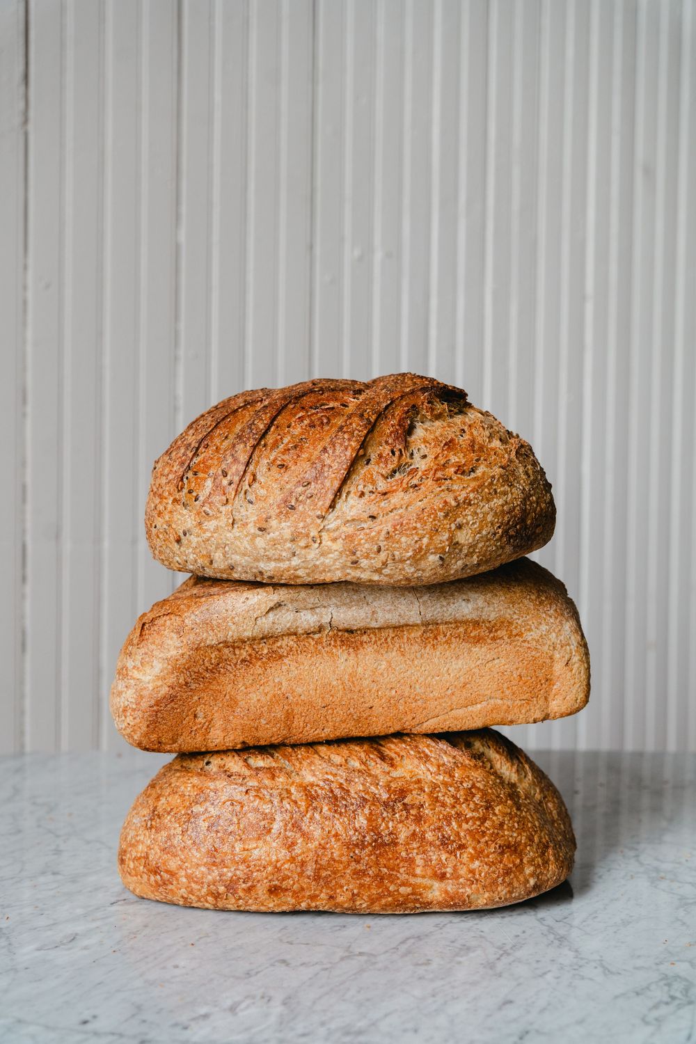 Three stacks of freshly baked artisanal sourdough bread loaves arranged on a light marble surface against a white background.