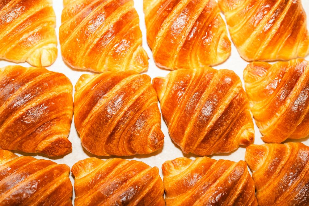 Golden brown croissants lined up in a row showing their flaky, layered texture against a light background.