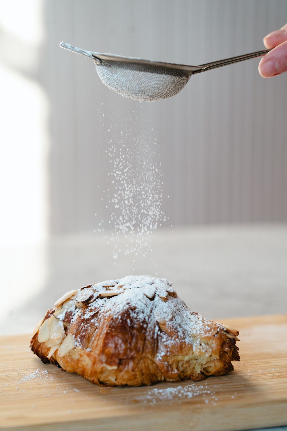 Powdered sugar being sifted from a metal strainer onto a freshly baked croissant on wooden cutting board.