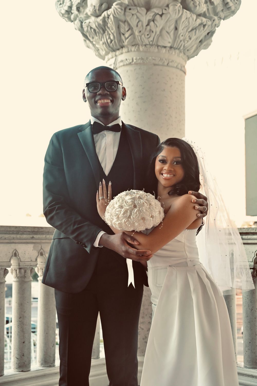 Smiling newlyweds posing with white rose bouquet by ornate column, groom in tuxedo, bride in white.