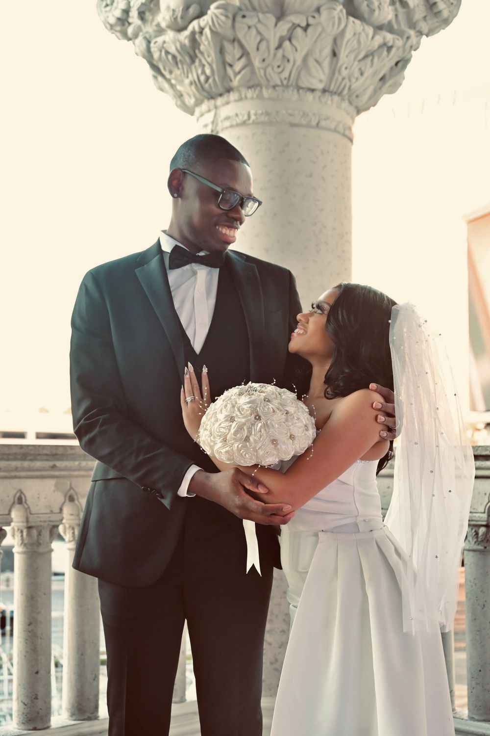 Happy newlywed couple sharing a smile beside ornate column, bride holding white floral bouquet.