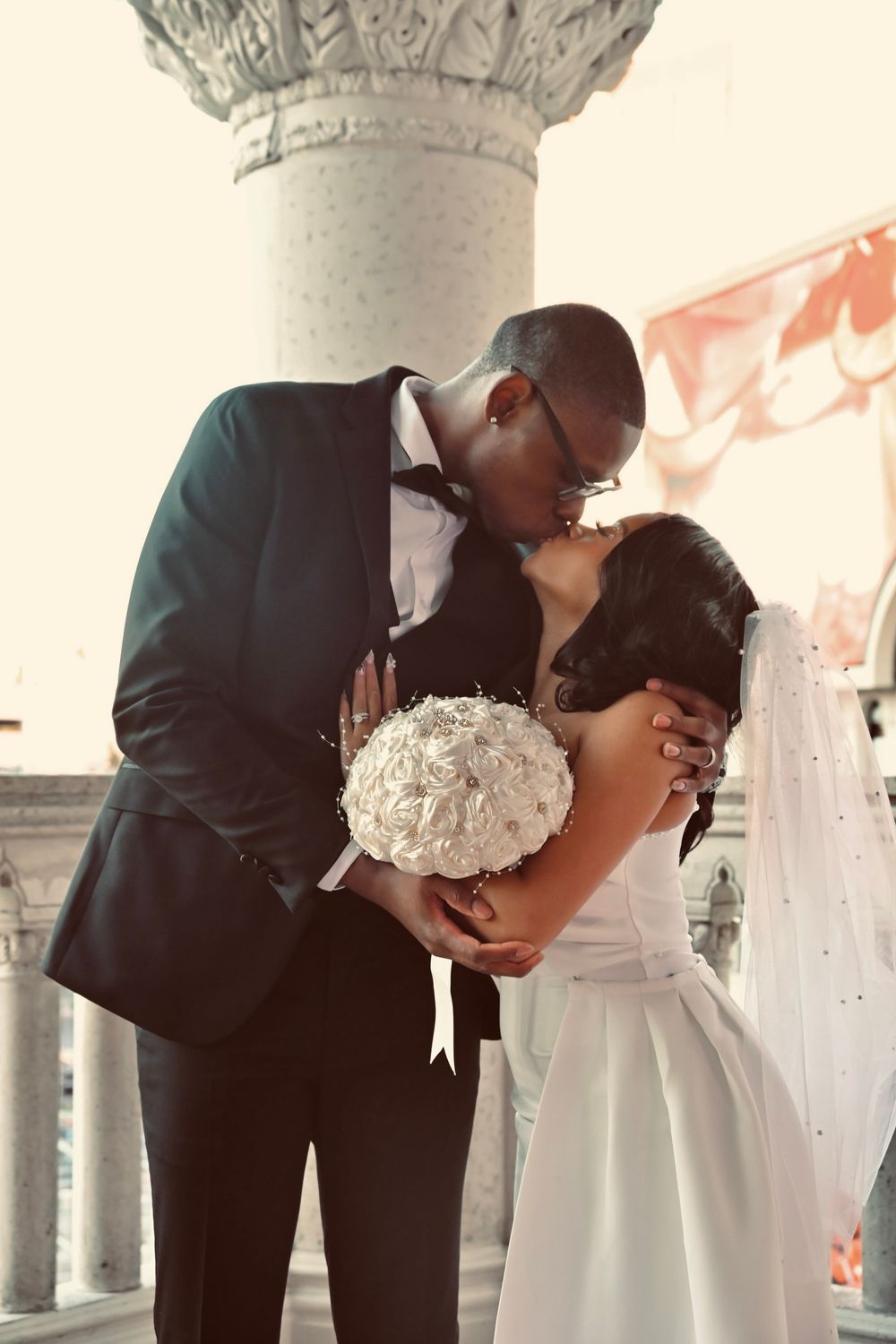 Groom in black tuxedo kissing bride holding white rose bouquet beside decorative stone column.
