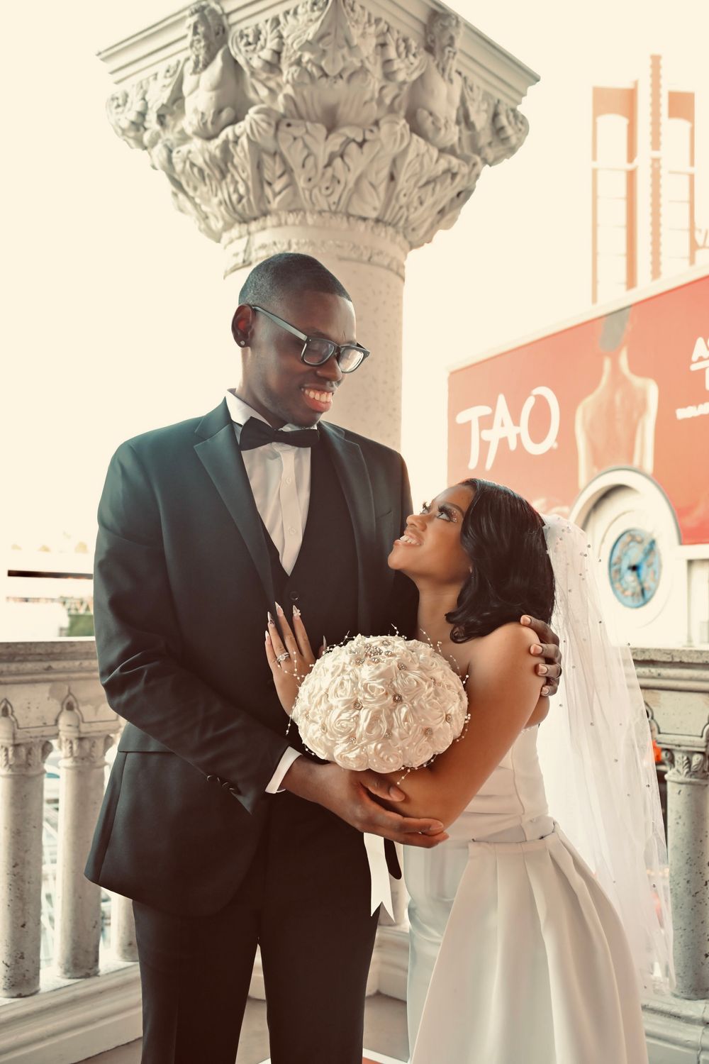 Elegant newlyweds posing with white bouquet near stone column with TAO sign in background.