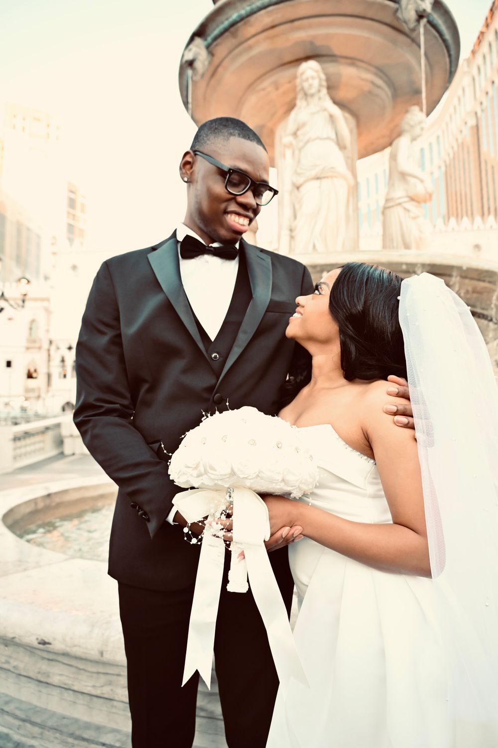 Groom in black tuxedo smiling at bride holding white bouquet by Las Vegas fountain.
