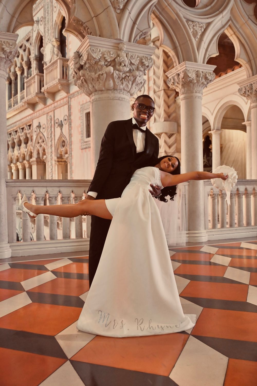 Groom dips bride in white gown reading Mrs. Rhyme on elegant Venetian-inspired marble floor.