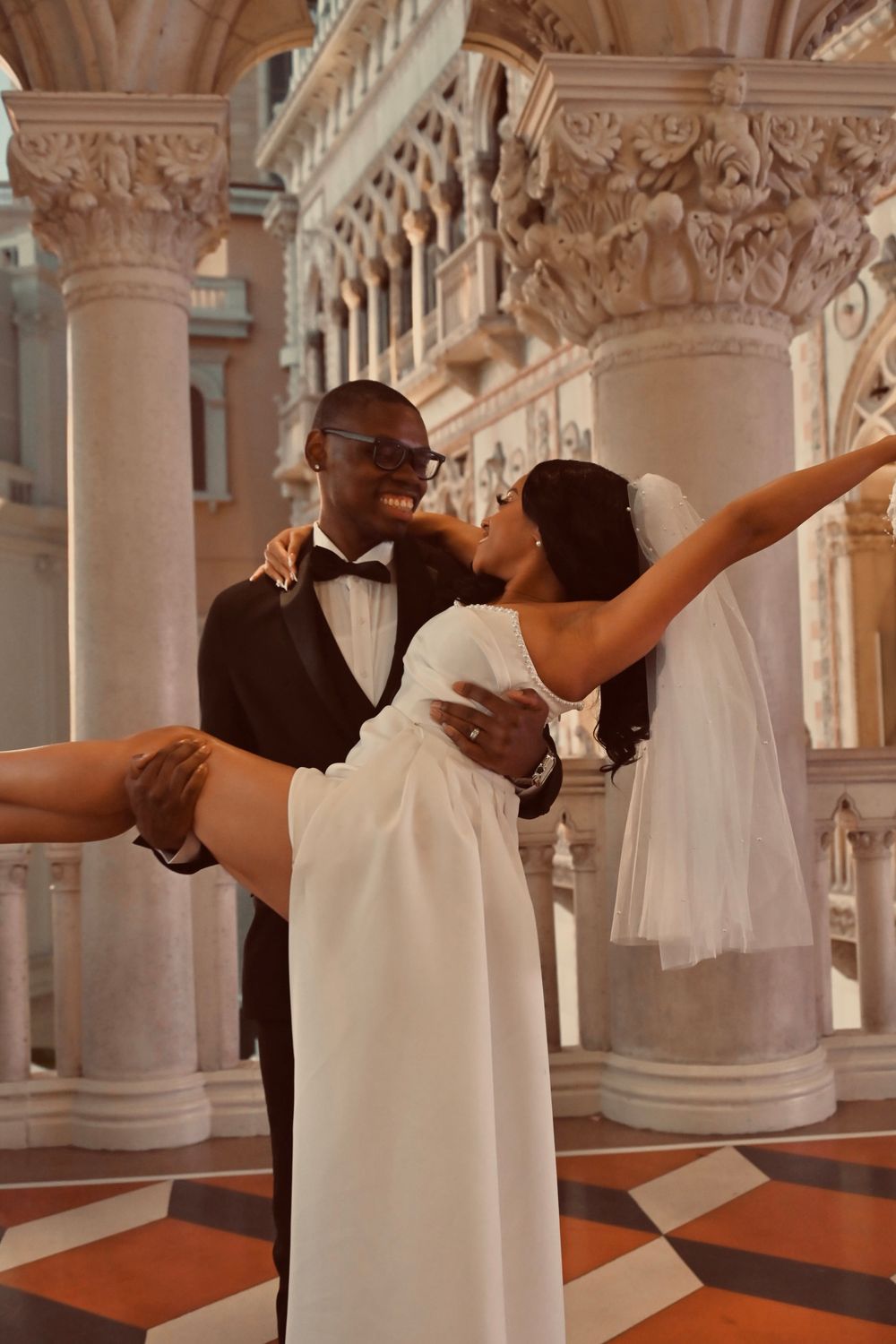 Smiling groom in tuxedo lifts bride in white wedding gown inside The Venetian Las Vegas.