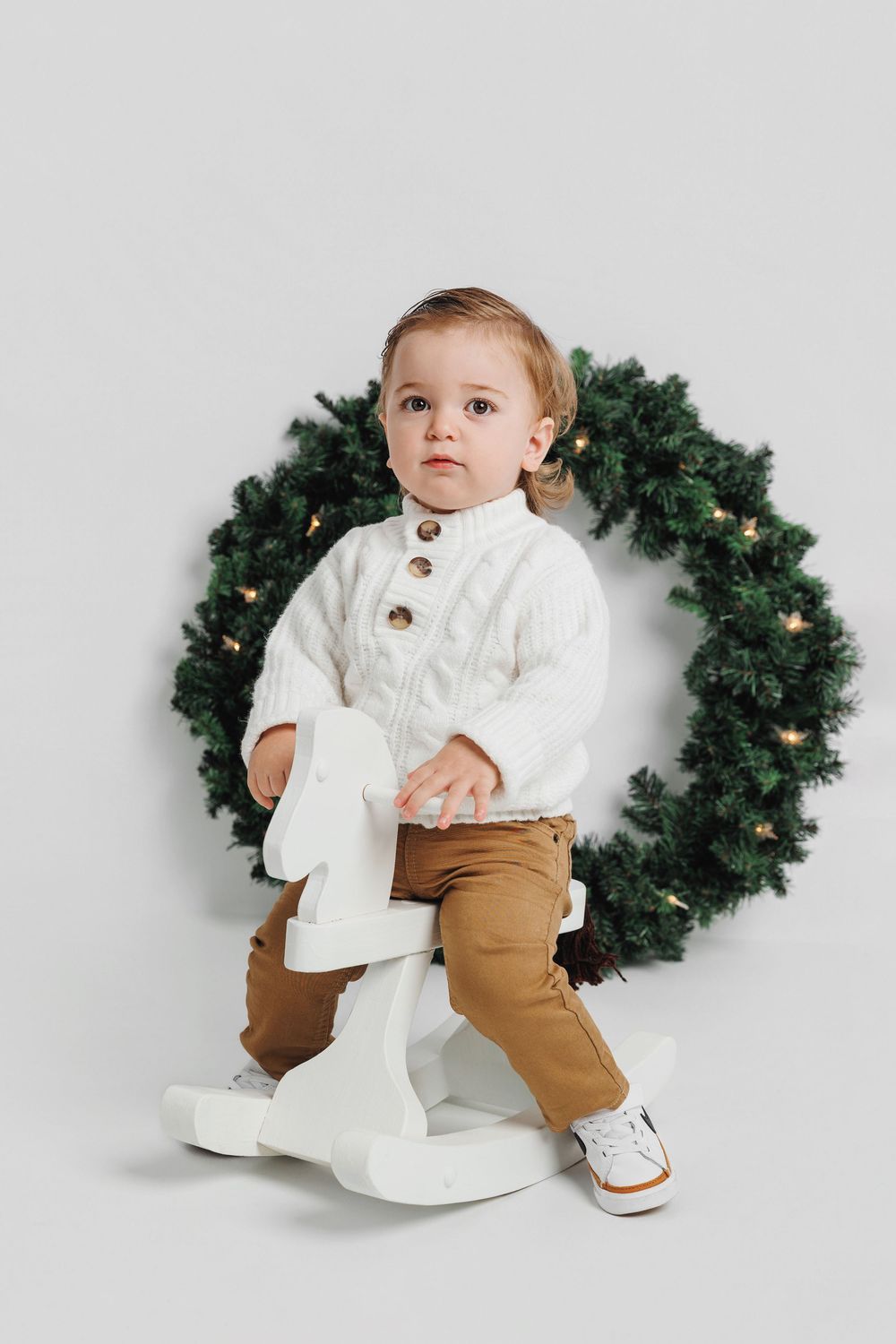 Baby in white sweater and brown pants poses with evergreen Christmas wreath against white backdrop.