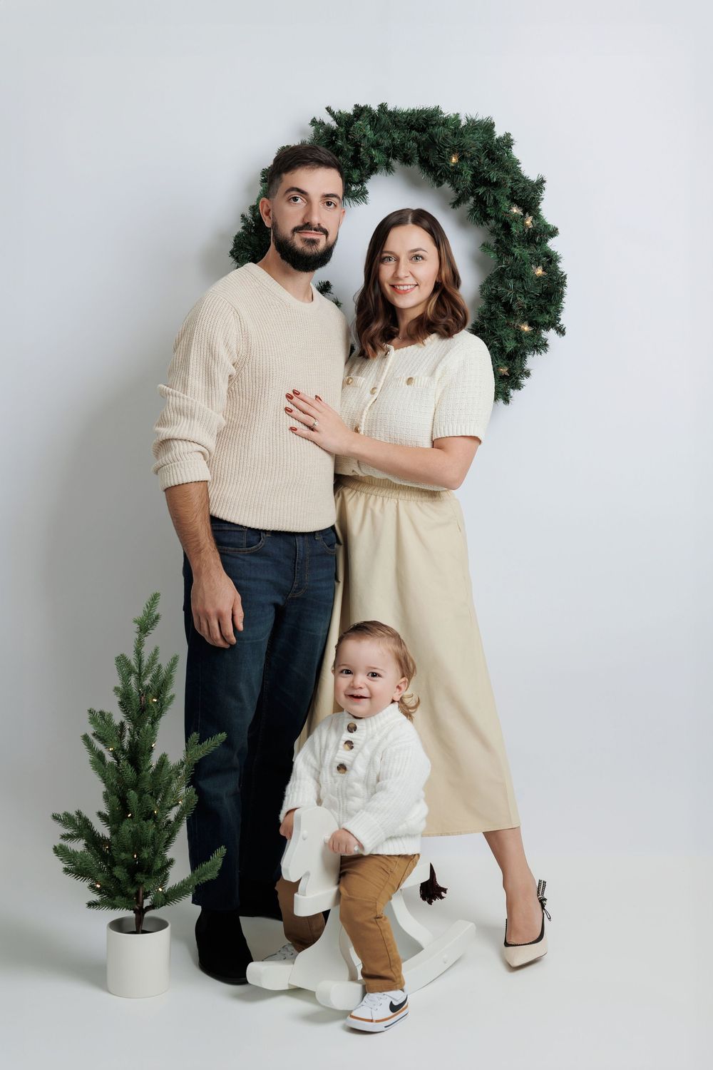 Family wearing coordinated cream and beige outfits poses against white backdrop with holiday greenery and small tree.