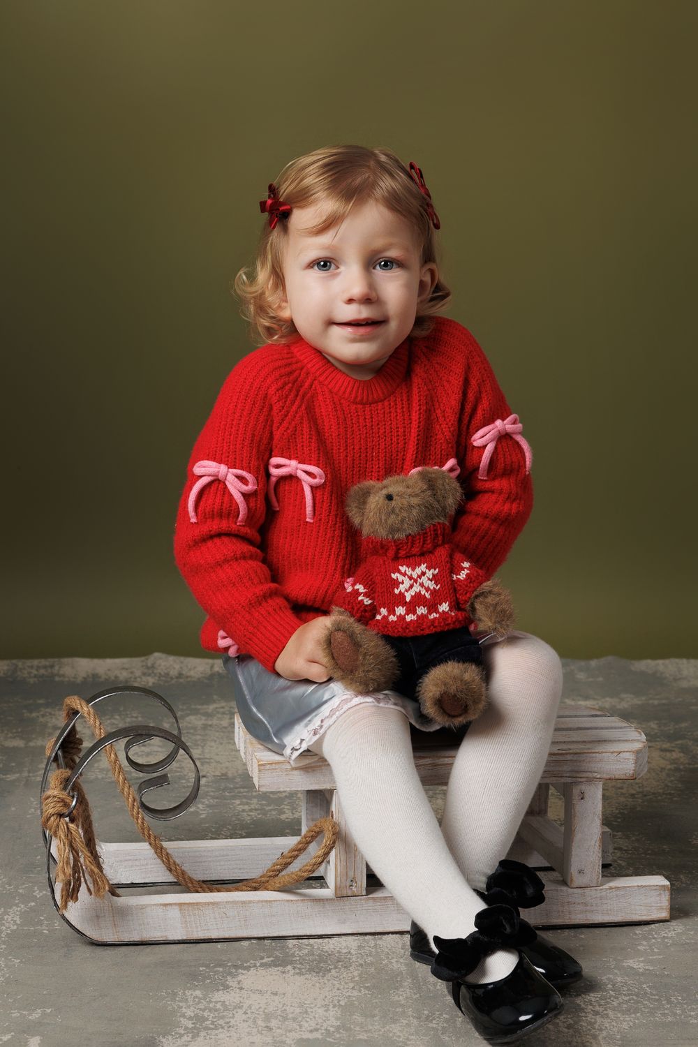 Small child wearing a festive red sweater sits on vintage wooden sled with white tights and black shoes.