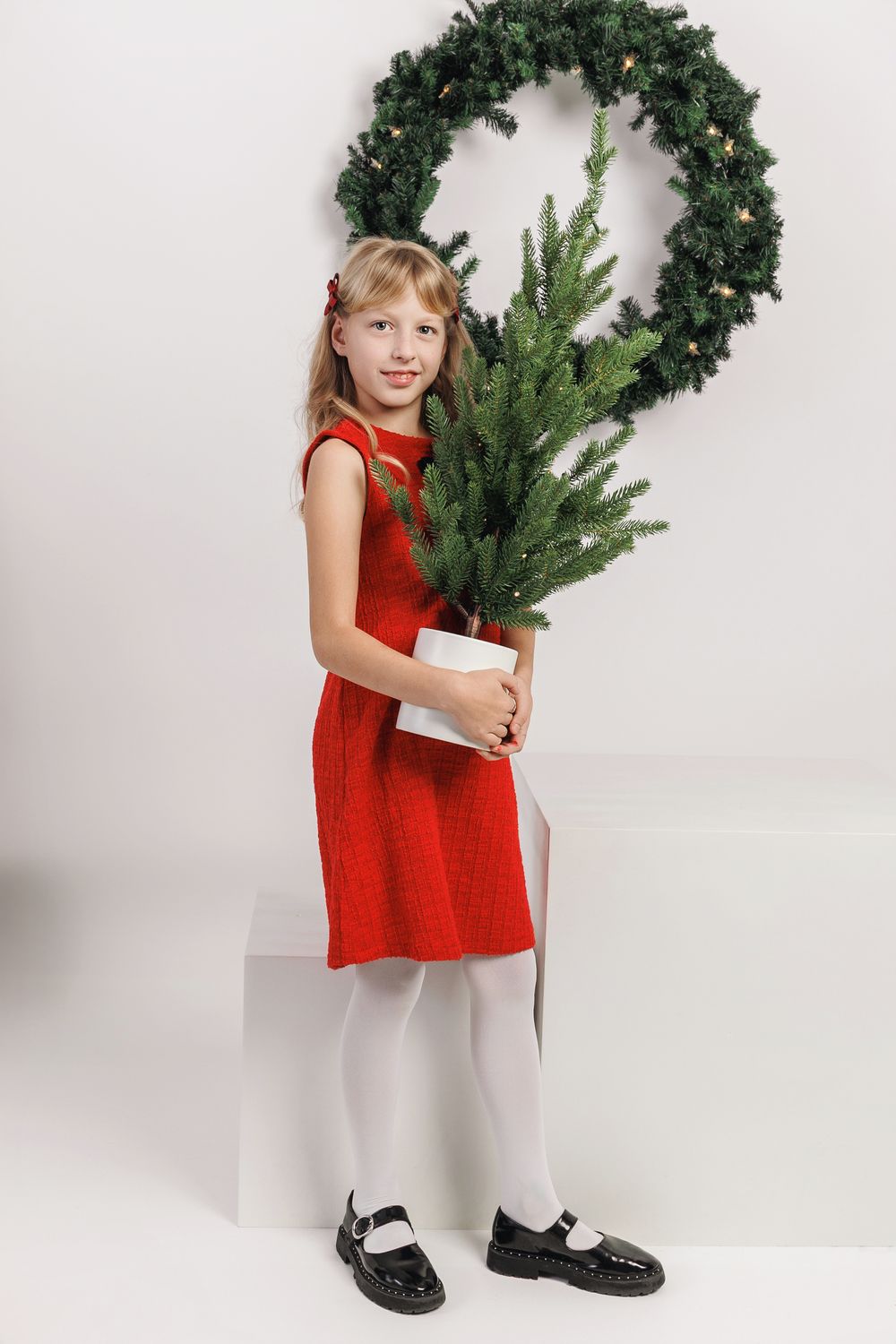 Child in red holiday dress holds evergreen branches and wreath while standing against white backdrop.