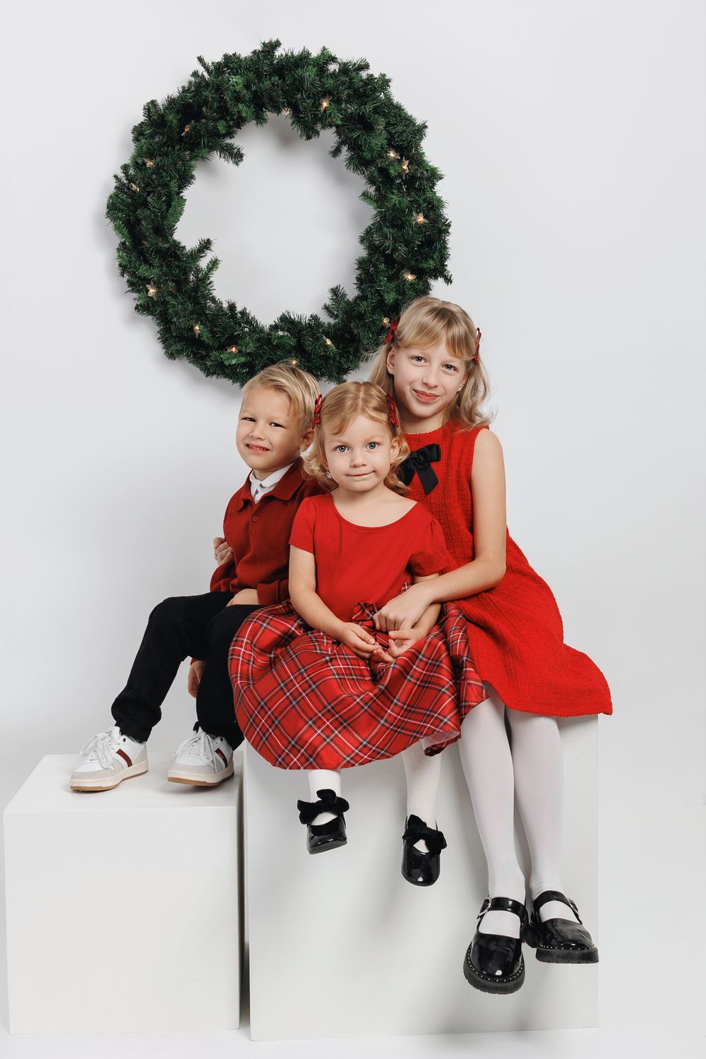 Three children in festive red holiday outfits sit together against a white backdrop with evergreen wreaths.
