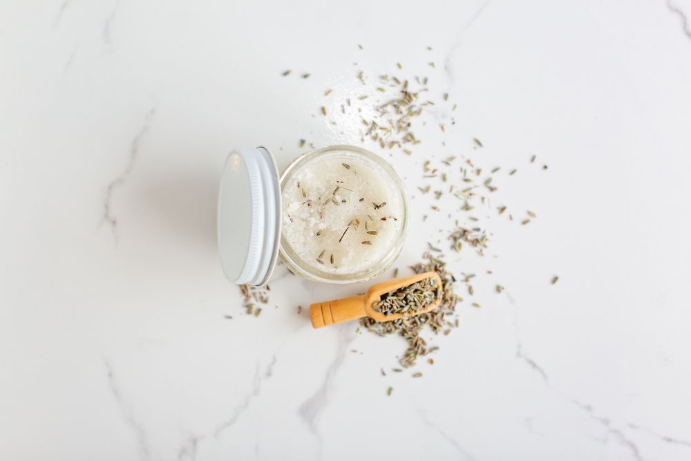 Natural bath salts scattered around glass jar on white marble backdrop.
