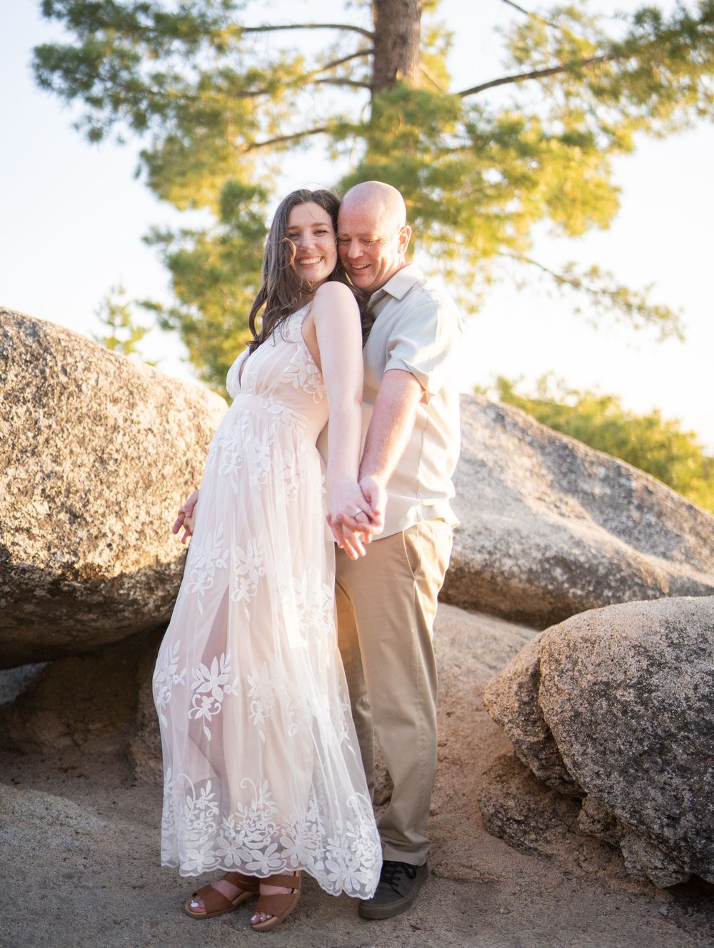 A couple poses together on rocky terrain at sunset with pine trees in the background.