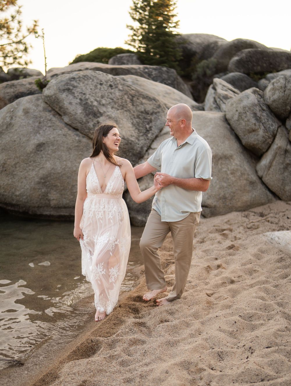 A couple shares a joyful moment walking along a sandy beach with large boulders in the background.