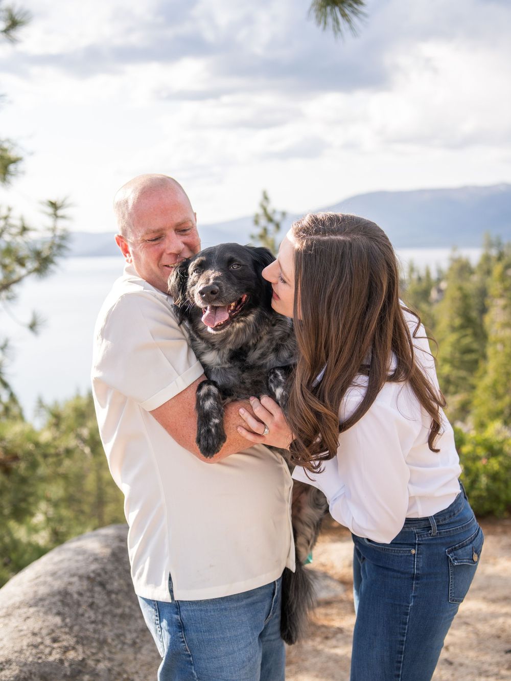 A couple and their happy black dog pose together outdoors in a scenic mountain setting with pine trees and rocks.