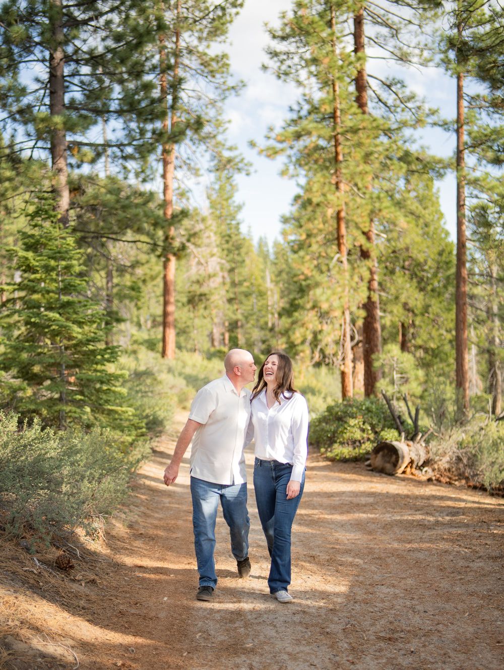 A couple walks hand in hand along a winding dirt path through a serene pine forest on a sunny afternoon.