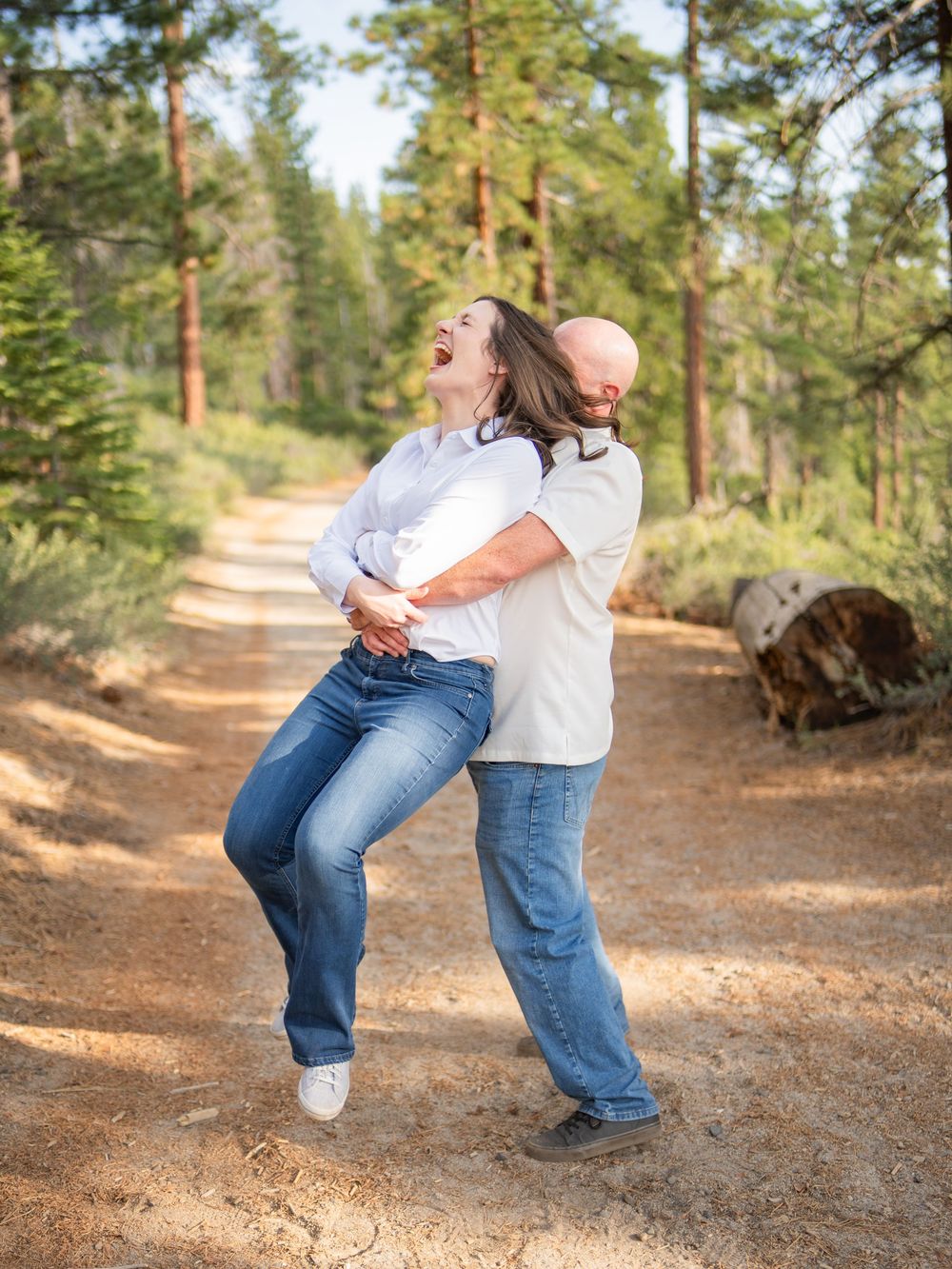 A joyful couple embraces and laughs together on a sunny forest trail surrounded by tall pine trees.