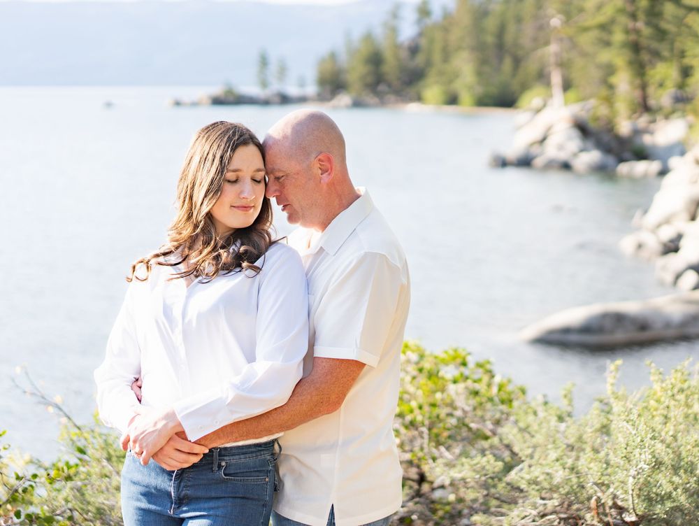 A couple embraces during an outdoor photoshoot near a scenic lake surrounded by evergreen trees and boulders.
