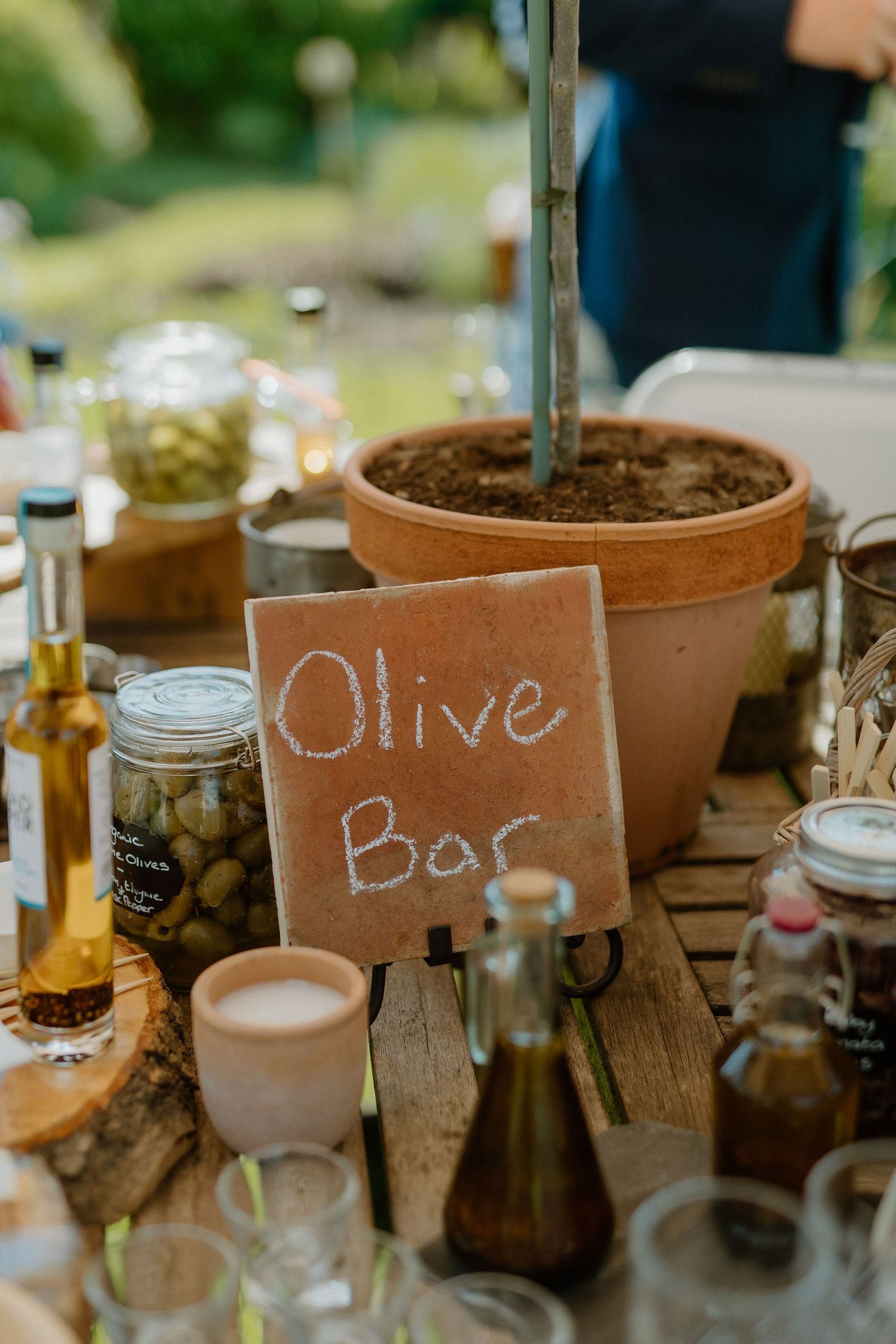 A rustic olive bar table setting with mason jars, bottles and a terracotta pot marked with a chalk sign.