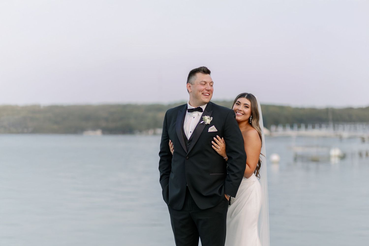A couple in formal wedding attire poses together by a serene waterfront at dusk in Bay Pointe Inn.