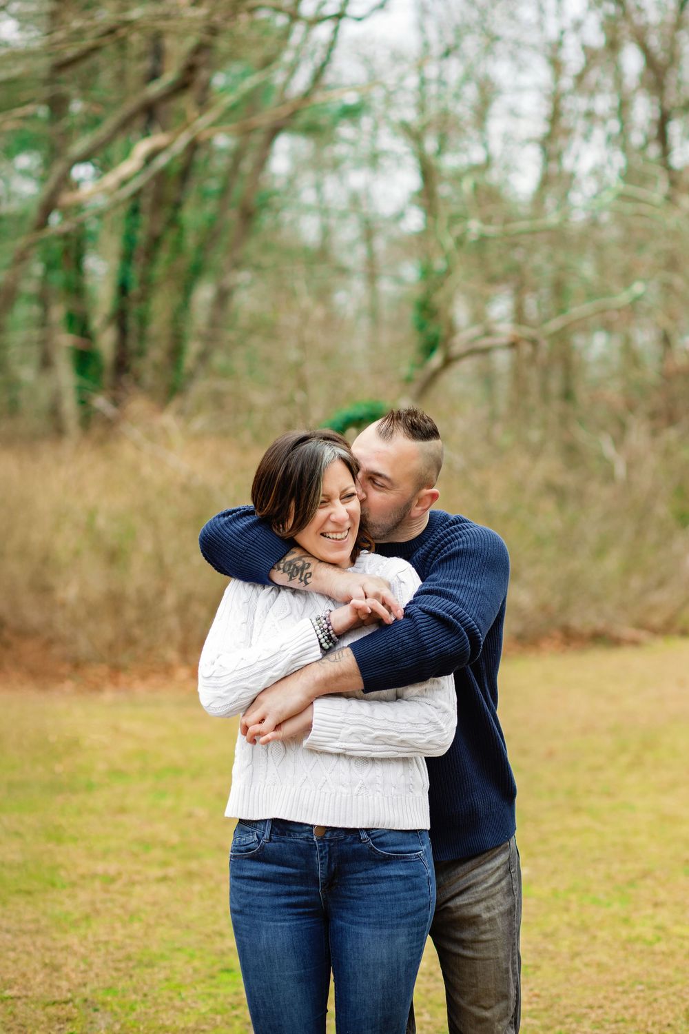 A couple shares playful candid moments together in a wooded outdoor setting wearing casual clothing.
