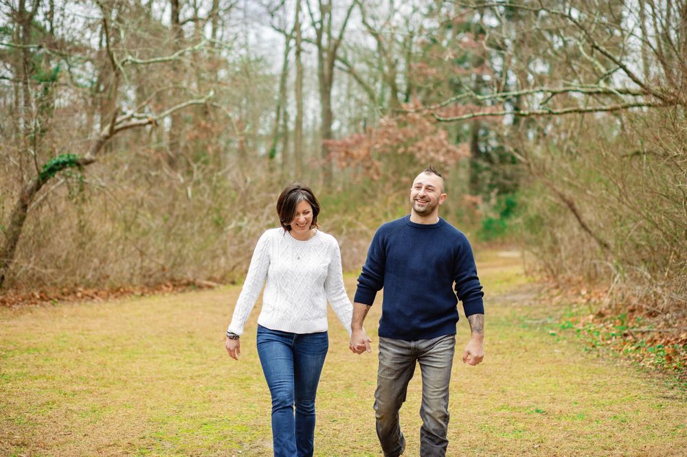 A couple walks hand in hand down a wooded trail during autumn with fallen leaves on the ground.