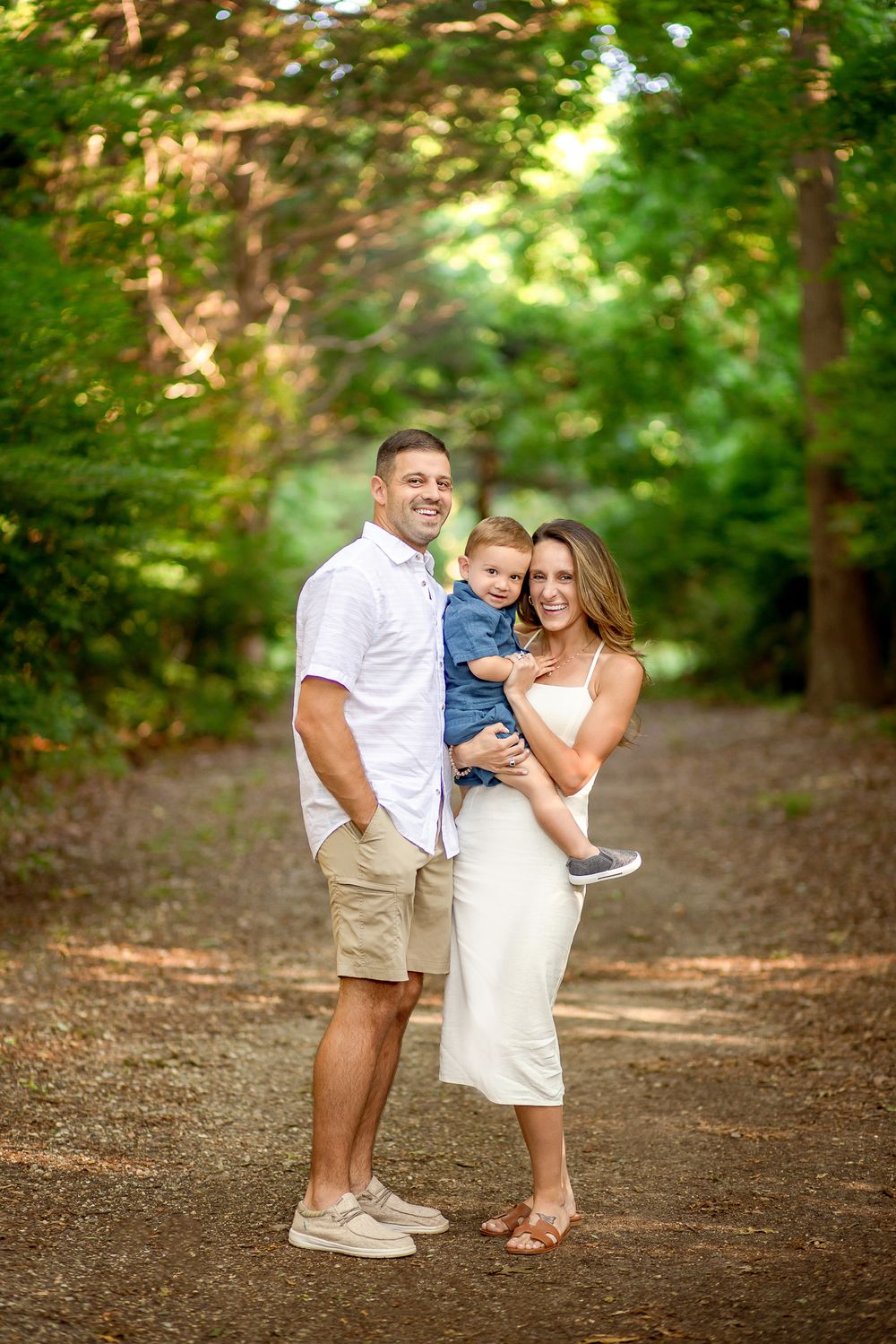 A family poses together on a tree-lined dirt path in a park during a sunny summer day.