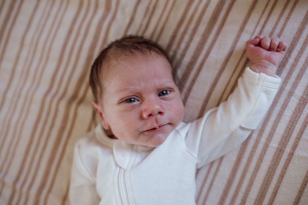 A baby in a white onesie lying on beige striped bedding with one arm raised upward. In home newborn session portsmouth, virginia