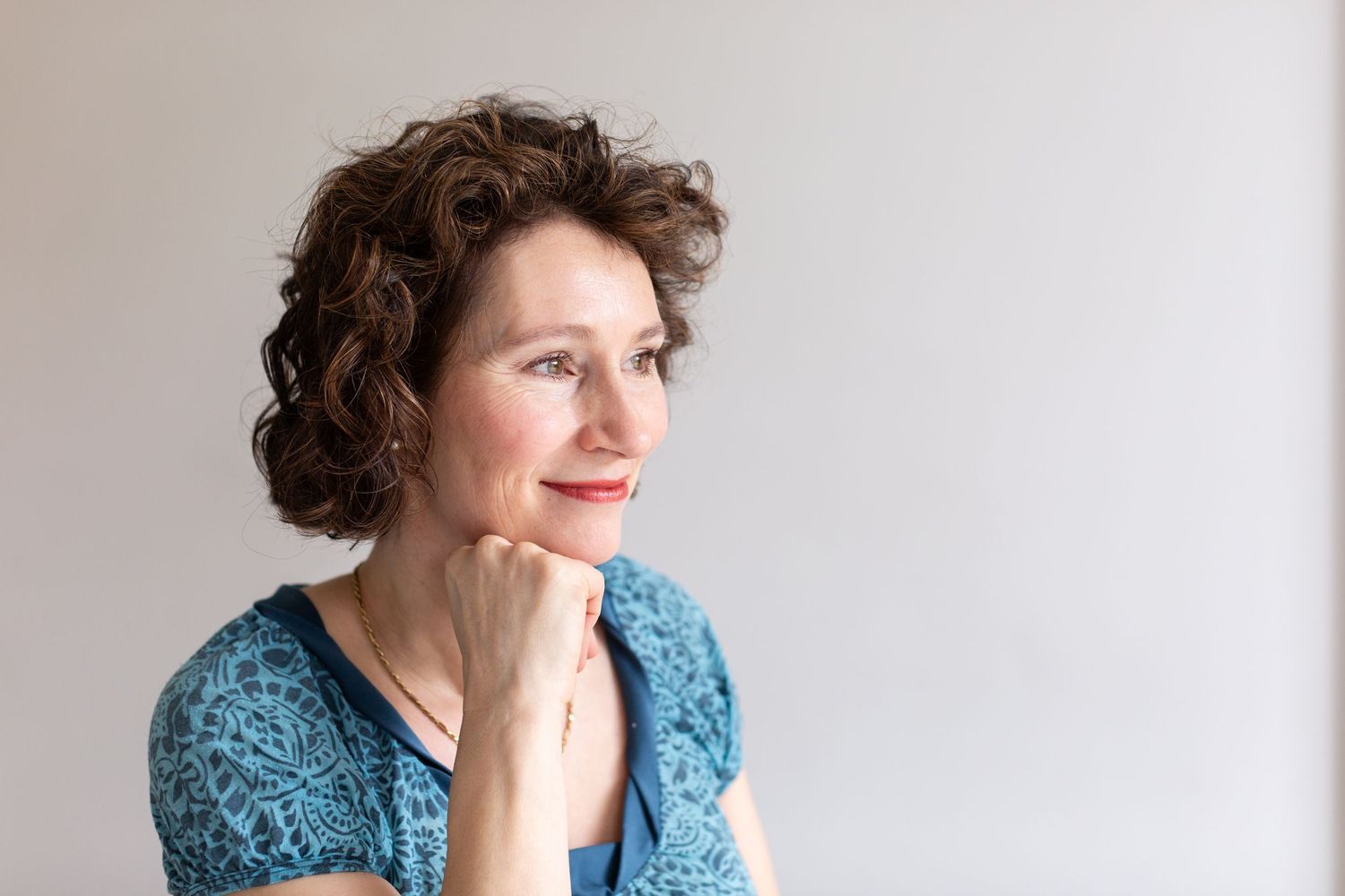 Person wearing a blue lace top rests chin thoughtfully on hand against light background.