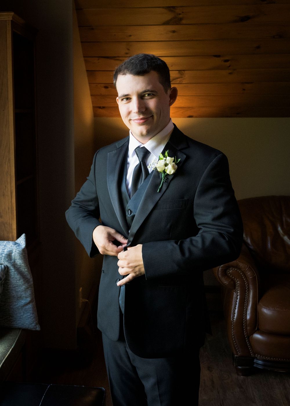 Groomsman in black suit adjusts his tie in a wood-paneled room.