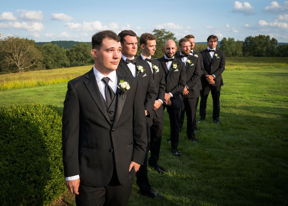 Groomsmen in black suits stand in a line on a grassy hill during a wedding.