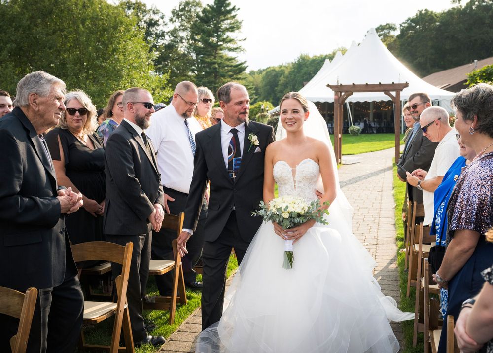 Outdoor wedding ceremony with guests seated on white chairs and a rustic wooden arch on a sunny day.