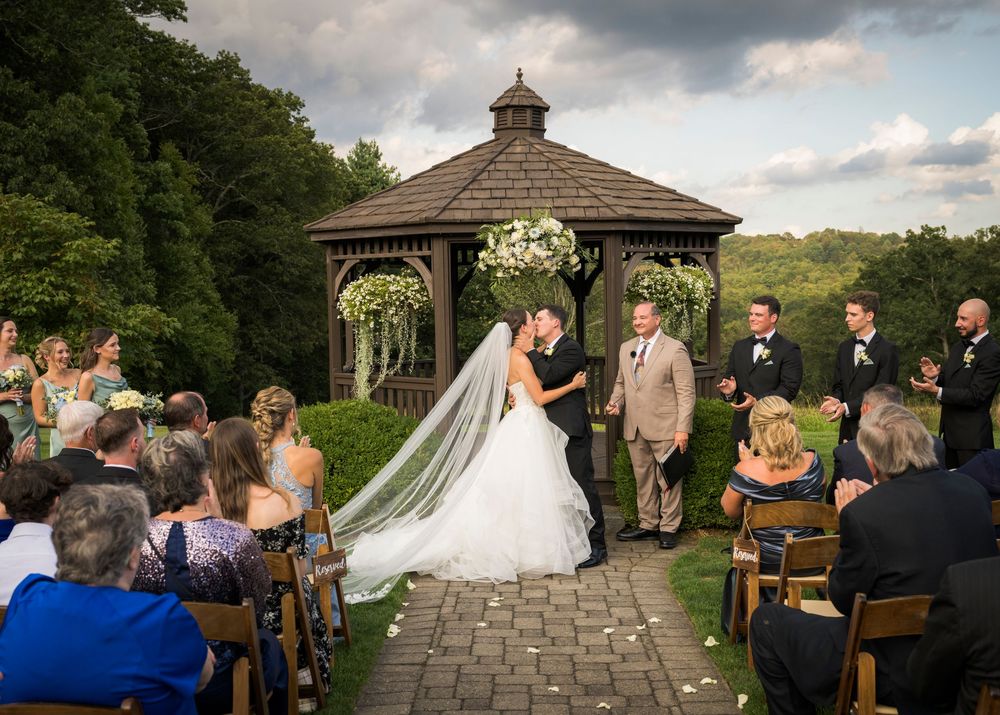 An outdoor wedding ceremony takes place under a wooden gazebo decorated with white flowers.