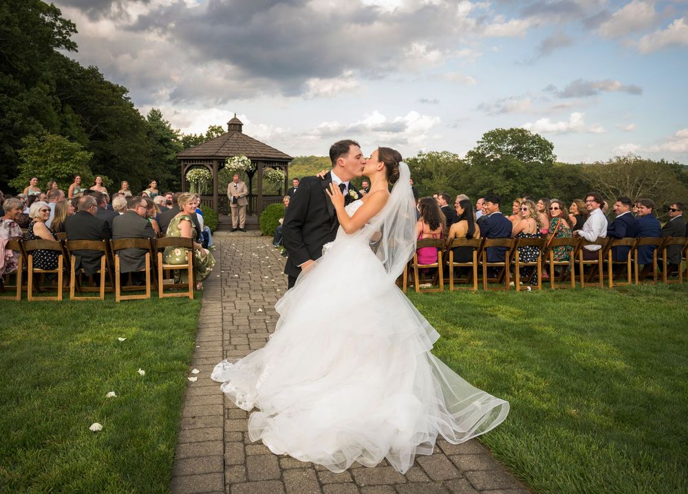Wedding ceremony in progress at an outdoor gazebo with guests seated on wooden chairs.