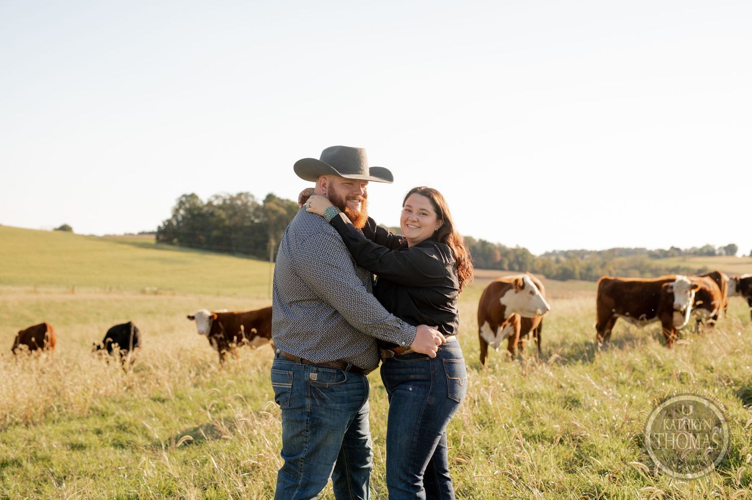 Couple sharing a tender moment in a sunny pasture with grazing cattle in the background.