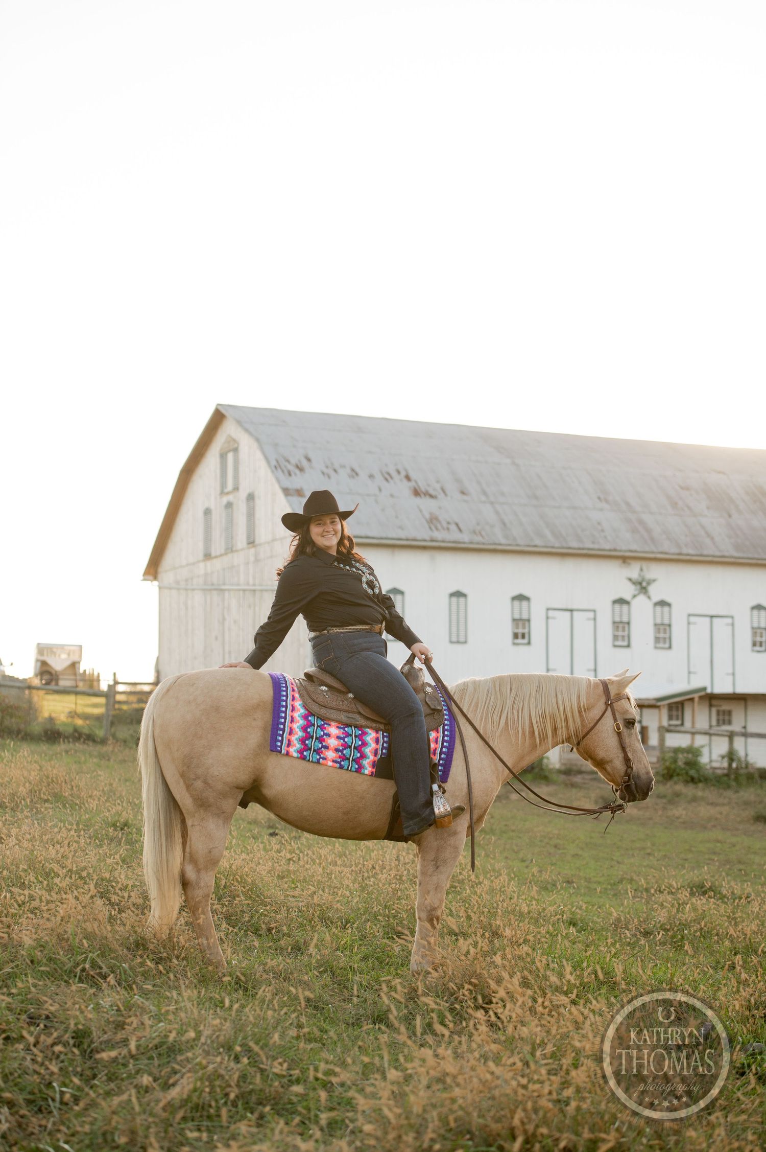 Rider on pale horse with colorful saddle blanket near white barn at sunset.