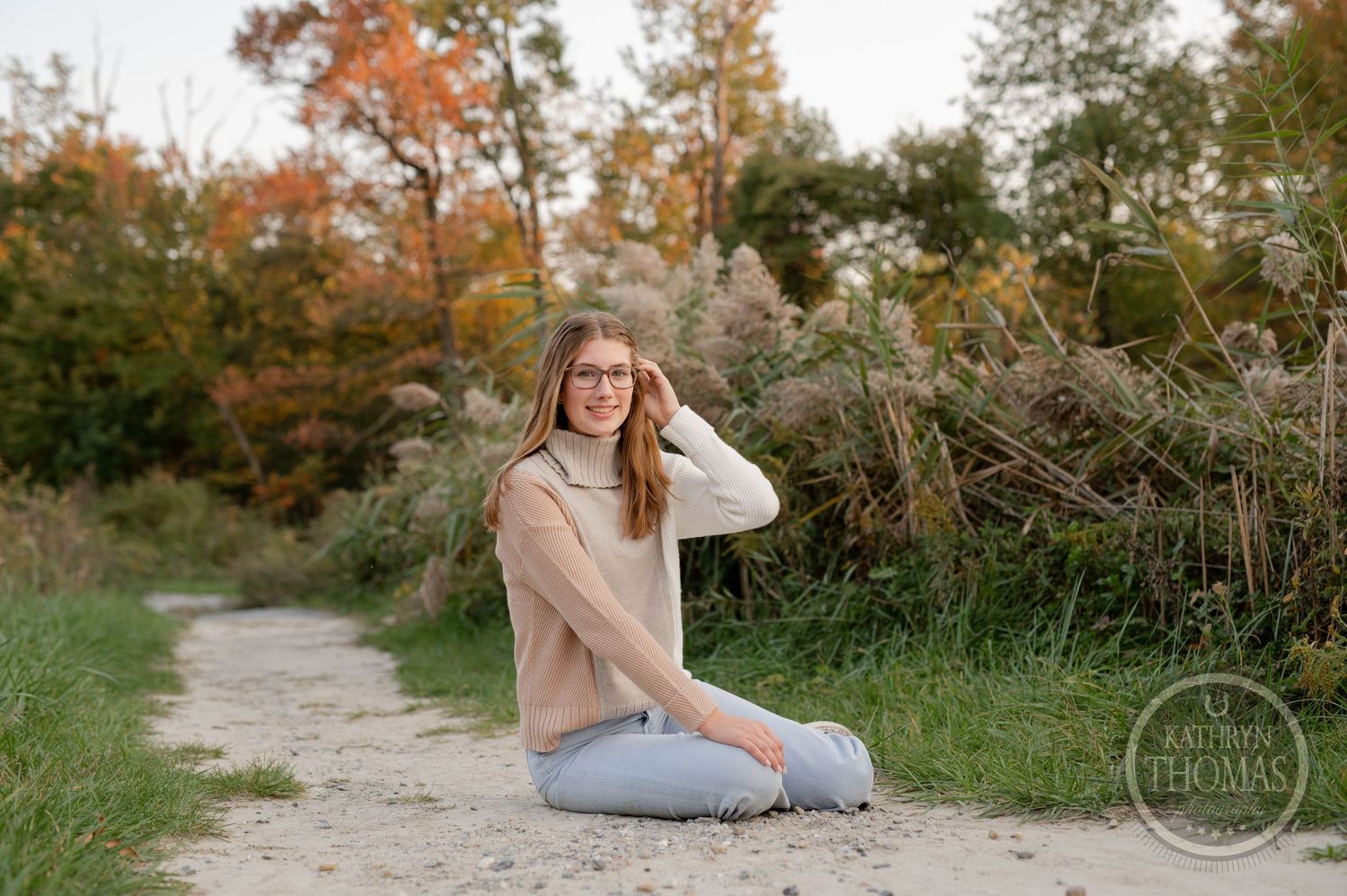 Outdoor portrait in beige sweater and jeans sitting on path with autumn foliage.