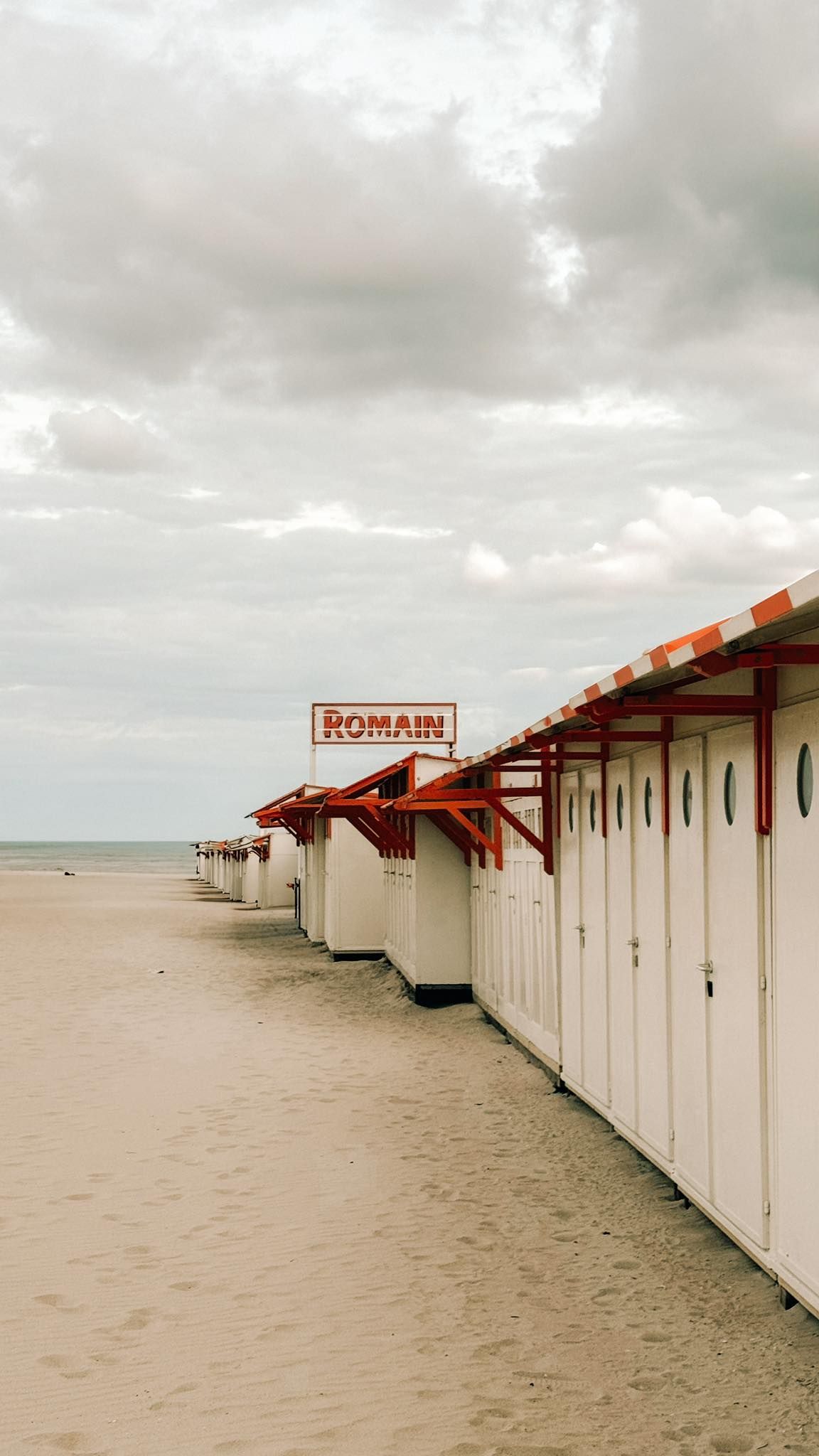 White beach cabanas with red striped roofs line the sandy shore under cloudy skies at a coastal resort beach.