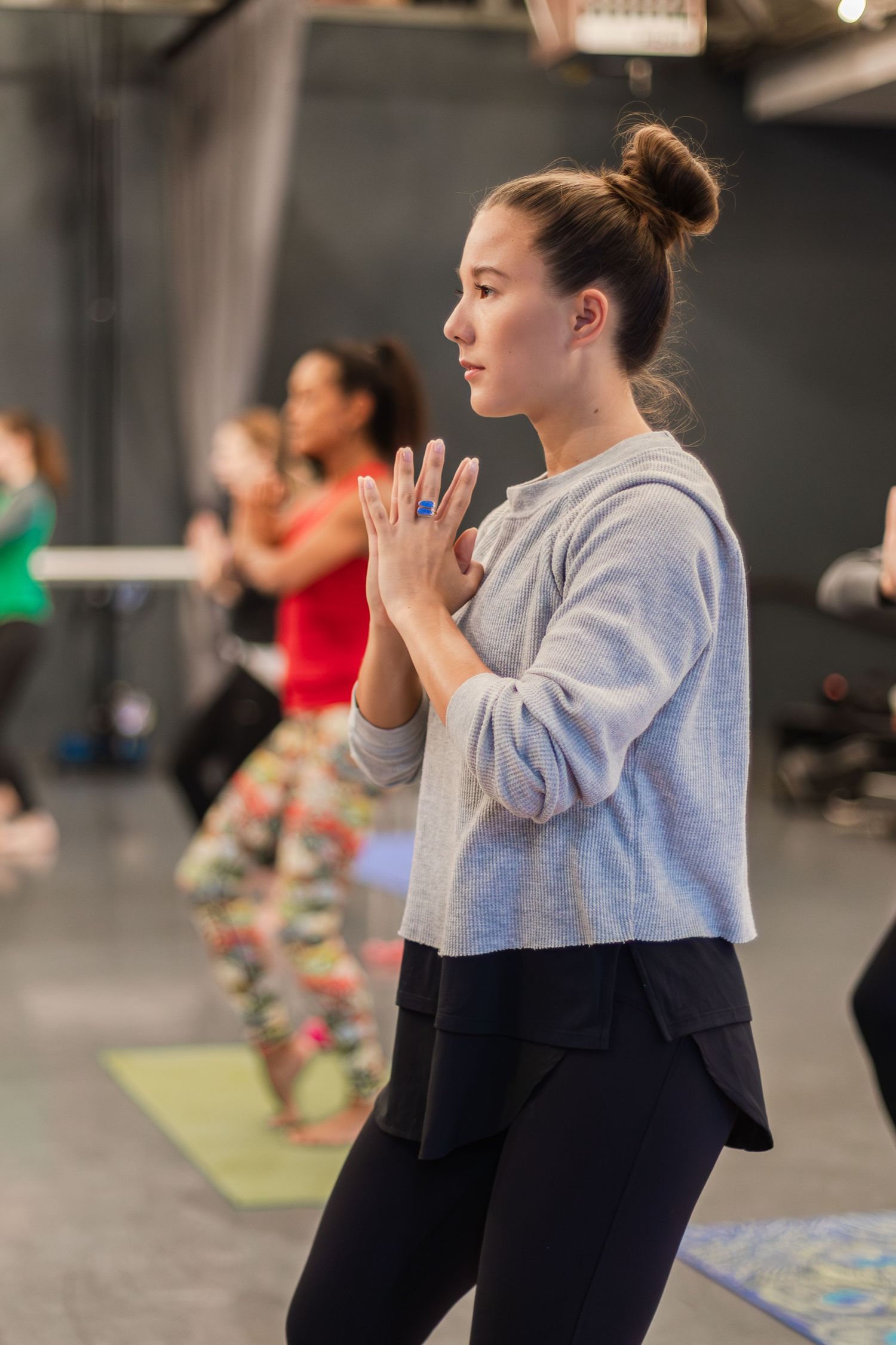 A person in a grey sweatshirt and black leggings practices yoga or meditation in a fitness studio with others visible behind.