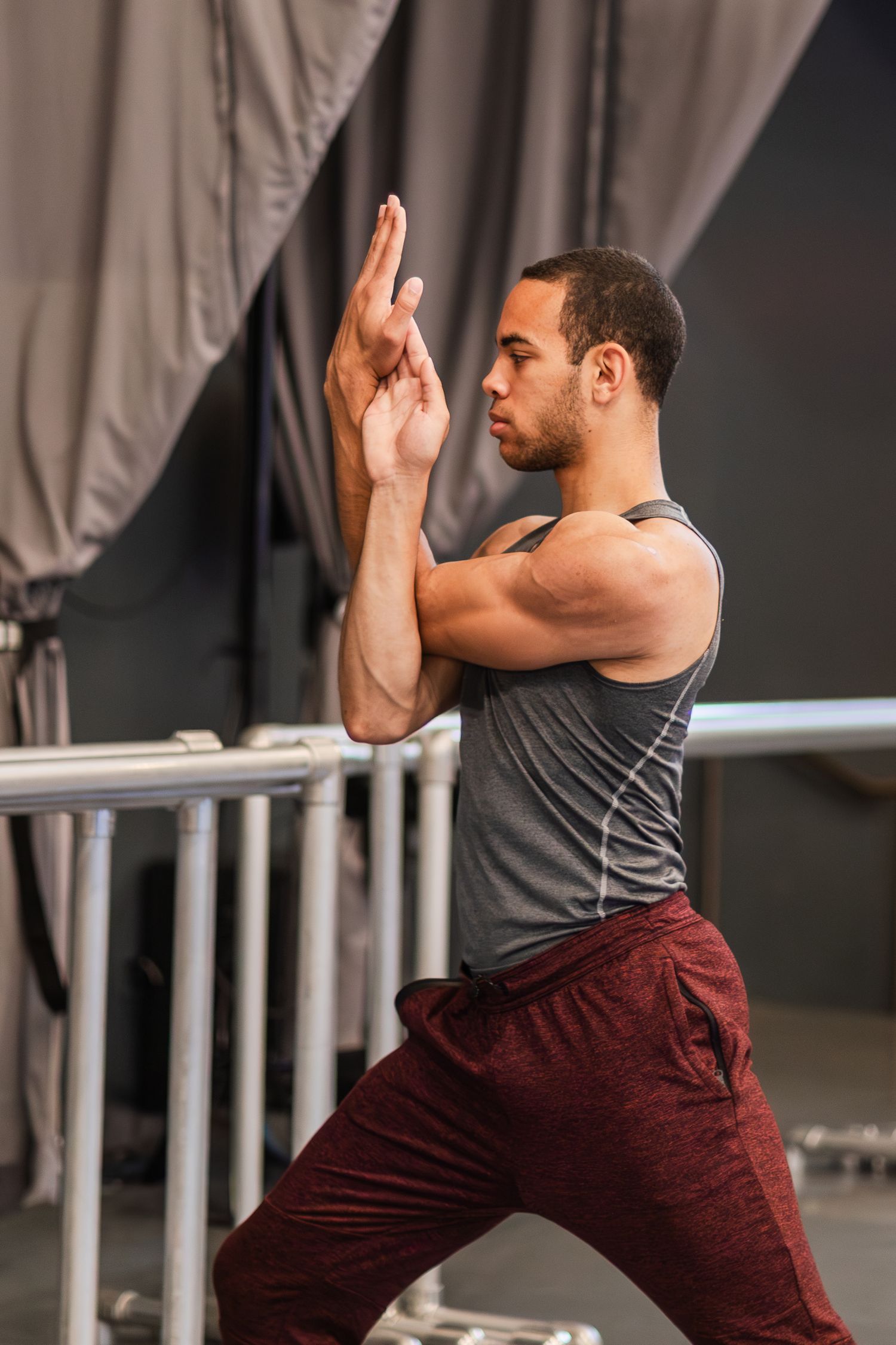 Dancer in gray tank top and burgundy pants stretches in profile against studio backdrop during rehearsal.